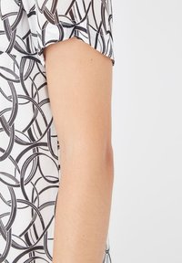 Brazo con una camisa de manga corta blanca con un patrón de líneas circulares negras, mostrado contra un fondo claro y liso.