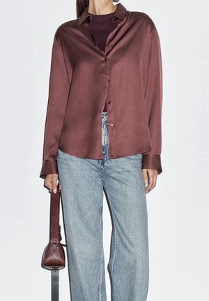 Vrouw draagt een glanzend bordeauxrood bloesje met knopen over een donker bordeauxrood topje, lichtblauwe hoge taille jeans, en houdt een bijpassende bordeauxrode handtas vast.