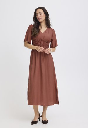 Frau trägt ein rostfarbenes Midikleid mit kurzen Ärmeln und seitlichen Schlitzen, steht mit sanft gefalteten Händen und schaut nach unten.