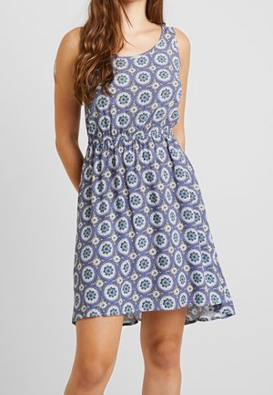 Mouwloze knielange jurk met blauw-wit cirkelvormig geometrisch patroon, nauw aansluitende taille, gedragen door een persoon met lang bruin haar, staand.