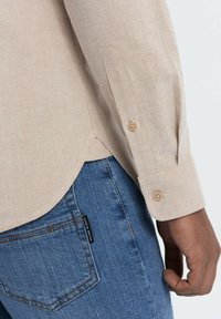 Beige langarmshirt met knoopmanchetten, gecombineerd met blauwe jeans. Het shirt heeft een zachte textuur en een gebogen zoom.