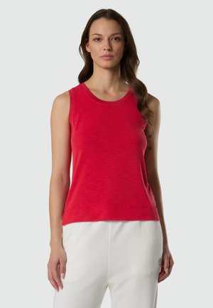 Donna con capelli lunghi castani, che indossa un top rosso senza maniche e pantaloni bianchi, in piedi davanti a uno sfondo grigio chiaro.