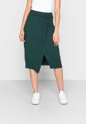 Kobieta ubrana w ciemnozieloną, kopertową spódnicę midi i białe trampki, stojąca na tle jednolitego białego tła.