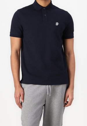 Homme portant un polo bleu marine ajusté avec une patte de boutonnage à deux boutons et un pantalon de survêtement gris avec cordon de serrage, debout devant un fond blanc.