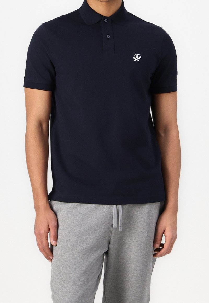 Homme portant un polo bleu marine ajusté avec une patte de boutonnage à deux boutons et un pantalon de survêtement gris avec cordon de serrage, debout devant un fond blanc.