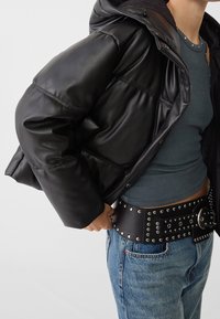 Chaqueta de plumón negra con capucha, acabado brillante y diseño acolchado. Llevada sobre una blusa en tono azul verdoso y combinada con un cinturón de cuero con tachuelas.