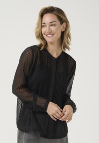 Schwarze Chiffonbluse mit vertikalen Falten, gepunkteter Textur und langen Ärmeln. Verfügt über einen runden Ausschnitt und einen kleinen goldenen Akzentknopf.