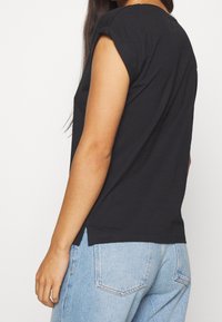 Camiseta de algodón negra con mangas cortas enrolladas, escote redondeado y aberturas laterales. La camiseta se combina con unos jeans de mezclilla azul claro.