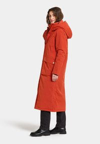 Long red winter coat with a hood, side pockets, and a textured collar. Paired with black pants and glossy ankle boots.