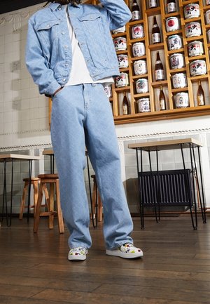 Osoba ubrana w jasnoniebieską kurtkę i spodnie dżinsowe oraz białe trampki, stojąca w pomieszczeniu z drewnianą podłogą i półkami z butelkami japońskiego sake.