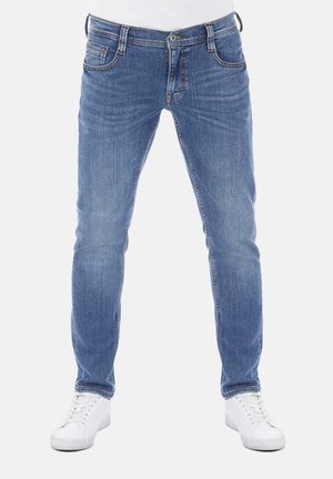 Blaue Slim-Fit-Jeans, getragen mit weißen Turnschuhen an einer Person, die vor einem weißen Hintergrund steht.