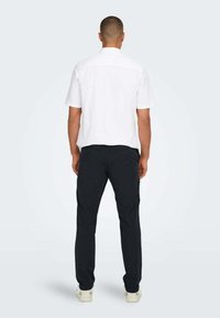 Camicia bianca a maniche corte con taglio dritto, abbinata a pantaloni scuri. Il tessuto sembra liscio, con colletto classico e senza motivi visibili.
