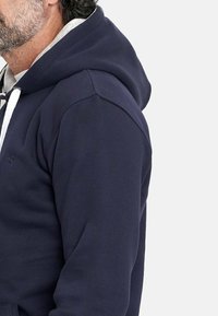 Sudadera con capucha de color navy, con un bolsillo tipo canguro, que cuenta con cordón y tejido suave y texturizado. Logo simple en el lado izquierdo del pecho.