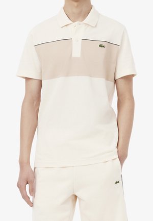 Mand iført en creme- og beige kortærmet poloshirt og matchende cremebukser med små grønne krokodillelogoer på begge.