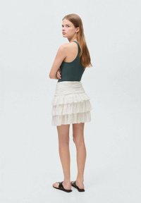 Donkergroen geribbelde tanktop, witte gelaagde mini-rok met ruffles en zwarte platte sandalen, gezien vanuit de achterkant. Glad materiaal en casual ontwerp.