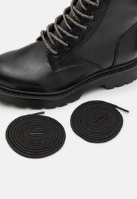 Bottines en cuir noir avec des boucles métalliques pour les lacets, semelle épaisse, et deux lacets ronds noirs posés à plat à côté. Texture lisse avec une légère brillance.
