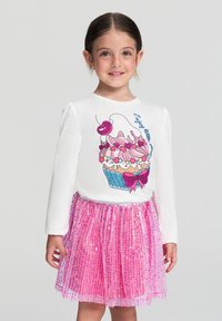 Młoda dziewczynka uśmiechająca się, ubrana w białą bluzkę z długim rękawem z grafiką babeczki oraz błyszcząca różowa spódnica z cekinami.