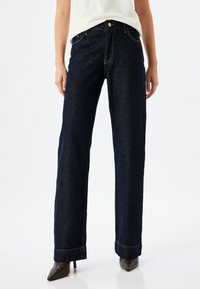 Jeans in denim blu scuro con fitting rilassato, caratterizzati da vita alta, gambe dritte e cuciture visibili. L'orlo è leggermente risvoltato.