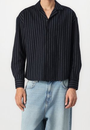 Homme portant une chemise noire à boutons avec des rayures verticales blanches et un jean bleu clair ample sur fond blanc.