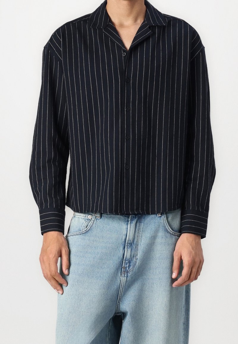 Homme portant une chemise noire à boutons avec des rayures verticales blanches et un jean bleu clair ample sur fond blanc.