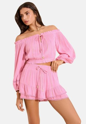 Frau trägt ein pinkfarbenes schulterfreies langärmeliges Crop-Top und passende hoch taillierte Shorts mit Rüschensaum und zarten Mustern.