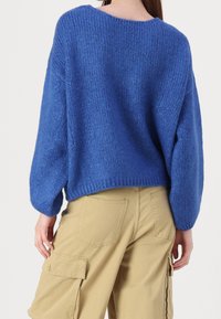 Blått stickat tröja med rundad hals, sänkta axlar och något kortare längd, kombinerat med beige cargobyxor. Texturerat tyg med lös passform.