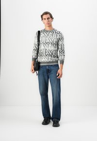 Uomo che indossa un maglione grigio con il testo "GUESS" ripetuto, jeans blu, scarpe nere e porta una borsa a tracolla nera su uno sfondo bianco semplice.
