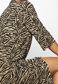 Un vestito beige con stampa zebra nera, caratterizzato da maniche a tre quarti e una vestibilità rilassata, con una texture evidente e un tessuto fluido.