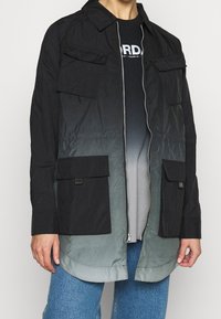 Giacca leggera nera con effetto sfumato che sfuma verso il grigio, dotata di chiusura con zip, due tasche a pattina e colletto a punta.