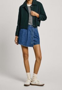 Ciemnozielona kurtka ściągana, biało-czarna koszula z długim rękawem w paski, dżinsowa spódnica typu wrap, białe trampki, beżowe skarpetki z teksturą prążków.