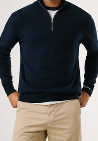 Pull en tricot bleu marine avec col à fermeture éclair partielle, poignets et ourlet côtelés, avec un logo sur le poignet. Porté sur une chemise blanche et un pantalon beige.