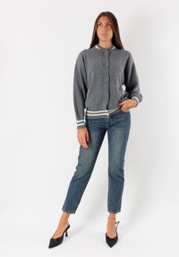 Donna in piedi contro uno sfondo bianco, che indossa un cardigan grigio, jeans blu e scarpe con tacco nere, con una mano in tasca e lo sguardo leggermente rivolto verso l'alto.