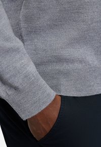 Maglione grigio in maglia con polsini a coste, indossato sopra un tessuto scuro, con una vista ravvicinata che mette in evidenza la trama e i dettagli della vestibilità.