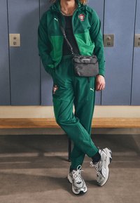 Grön sportdräkt med en svart Puma-emblem, bestående av en jacka med dragkedja och matchande byxor. Vita och svarta sneakers fulländar looken.