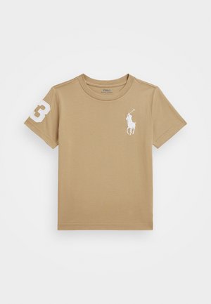 Bézs pamut póló, amelyen egy kis fehér logó található a mellrészen, valamint egy fehér "3" hímzett szám a bal ujjon. Kerek nyakú.