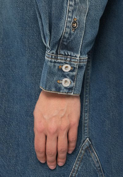 Μανσέτα πουκάμισου από τζιν σε ανοιχτό μπλε χρώμα με εμφανείς ραφές, που διαθέτει τρία ασημένια κουμπιά τύπου σνάπ και υφή που συμπληρώνει το ύφασμα.
