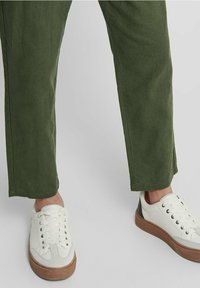 Olijfgroene, rechte stoffen broek met een gladde textuur. Gedragen met witte sneakers met een bruine rubberen zool en vetersluiting.