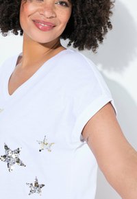 Bílé tričko s výstřihem do V, zdobené lesklými flitrovými vzory hvězd ve stříbrné a zlaté barvě. Krátké ohrnuté rukávy a měkká textilie.