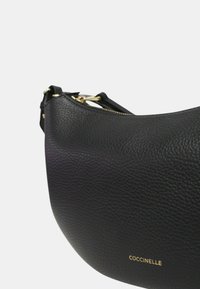 Borsa a mano in pelle nera con finitura testurizzata, forma curva, hardware dorato e zip visibile. Nome del marchio "Coccinelle" impresso in oro.