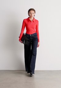 Tricou polo roșu cu mâneci lungi, guler și detaliu cu nasturi, purtat cu blugi larghi de culoare albastru închis. Modelul poartă o geantă plic neagră.