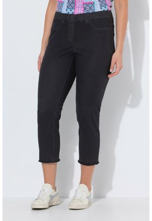 Kobieta ubrana w czarne skrócone jeansy z nieobrobionymi krawędziami, kolorową koszulę we wzory oraz białe trampki z jasnoniebieskimi akcentami.