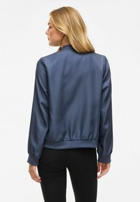 Giacca bomber in satin blu navy con polsini e orlo a coste, caratterizzata da un colletto alto e una texture liscia e riflettente. Jeans neri visibili sotto.