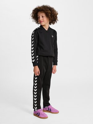 Enfant debout portant un survêtement noir avec des bandes blanches en chevron et des baskets roses, légèrement tourné vers la droite avec des cheveux bouclés.