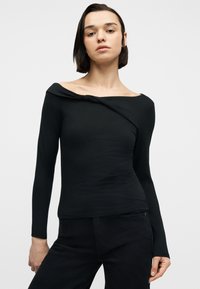 Νεαρό άτομο με κοντά σκούρα μαλλιά, φορώντας εφαρμοστό μαύρο τοπ με μακριά μανίκια που αφήνει τους ώμους ακάλυπτους και μαύρο παντελόνι, μπροστά σε απλό φόντο.