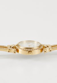 Montre analogique dorée avec un bracelet texturé, des accents de pierres précieuses, un cadran blanc et des index horaires dorés. La lunette et la couronne détaillées rehaussent le design.