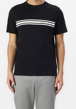 Mand iført sort t-shirt med korte ærmer med to hvide horisontale striber og teksten "Hilfiger Est 1985 New York" på brystet.
