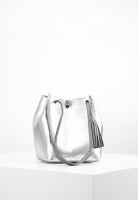 Sac seau métallique argenté avec une texture lisse, deux anses et un accent en frange. Sa forme est douce et arrondie.
