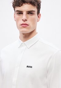 Biele polo tričko s textúrovanou tkaninou, s čiernym logom "BOSS" na ľavej strane hrudníka a klasickým golierom so zapínaním na gombíky.