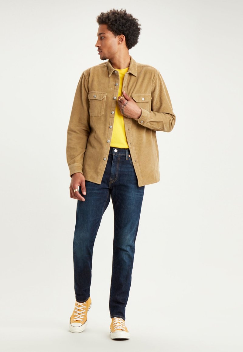 Chemise en velours côtelé beige avec deux poches poitrine, t-shirt jaune en dessous, jean bleu foncé et baskets oranges avec des lacets blancs.