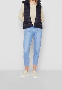 Marinblå puffer-väst med huva över en beige stickad tröja, i kombination med ljusblå uppvikta jeans och krämfärgade sneakers. Slät, vadderad textur.